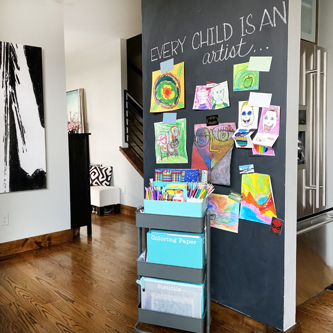 Rolling cart filled with art supplies in front of a chalkboard wall with colorful kids' artwork 